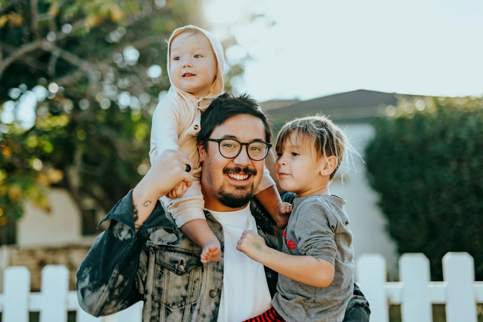 A smiling father carrying his two young children outside their home